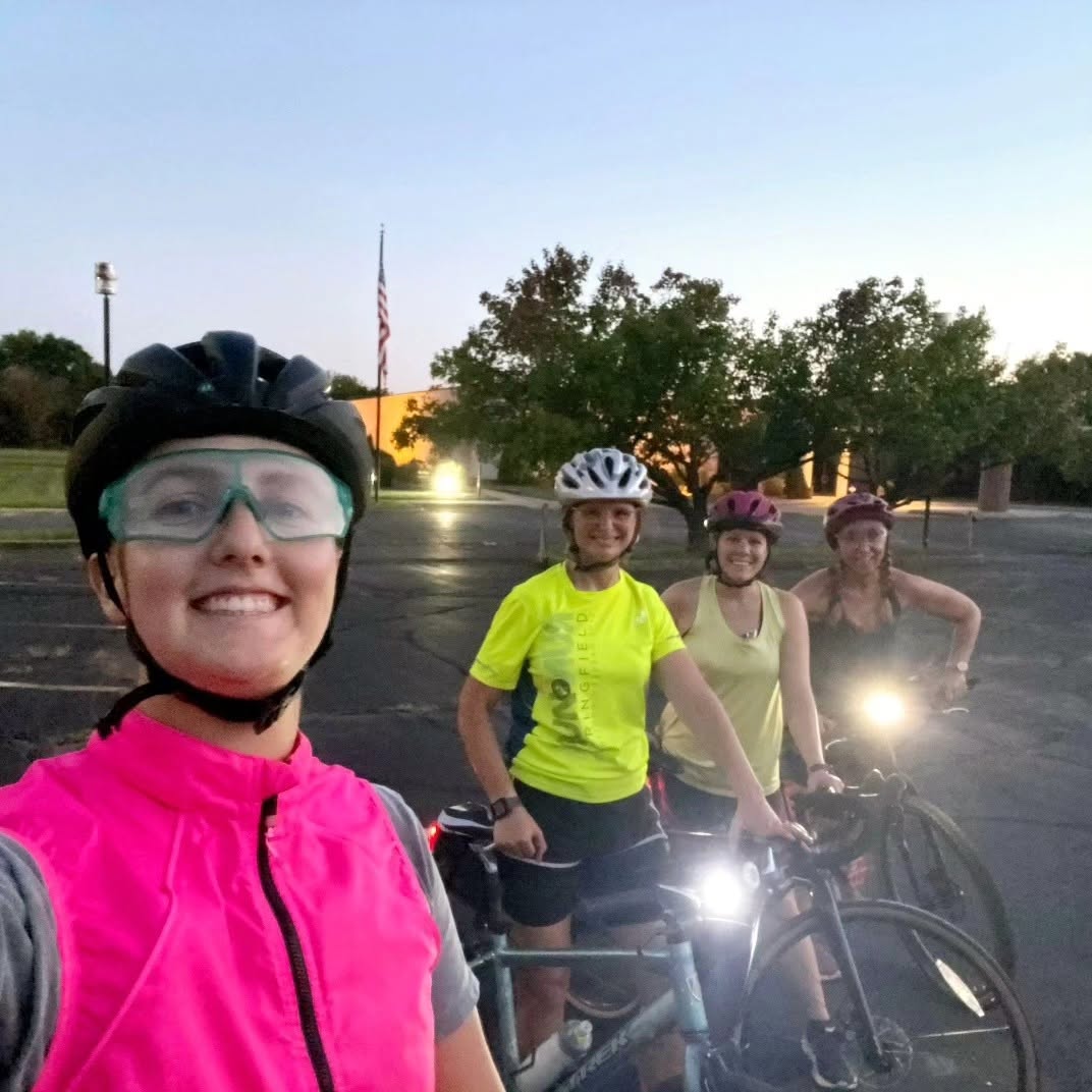 Four women cyclists in high-visibility gear smiling for a dusk selfie with bike lights on